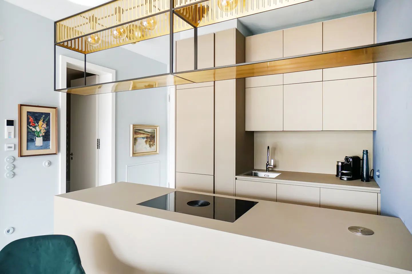 Modern kitchen with beige cabinets, a black cooktop on the island, and a gold light fixture. Paintings hang on the light blue walls.