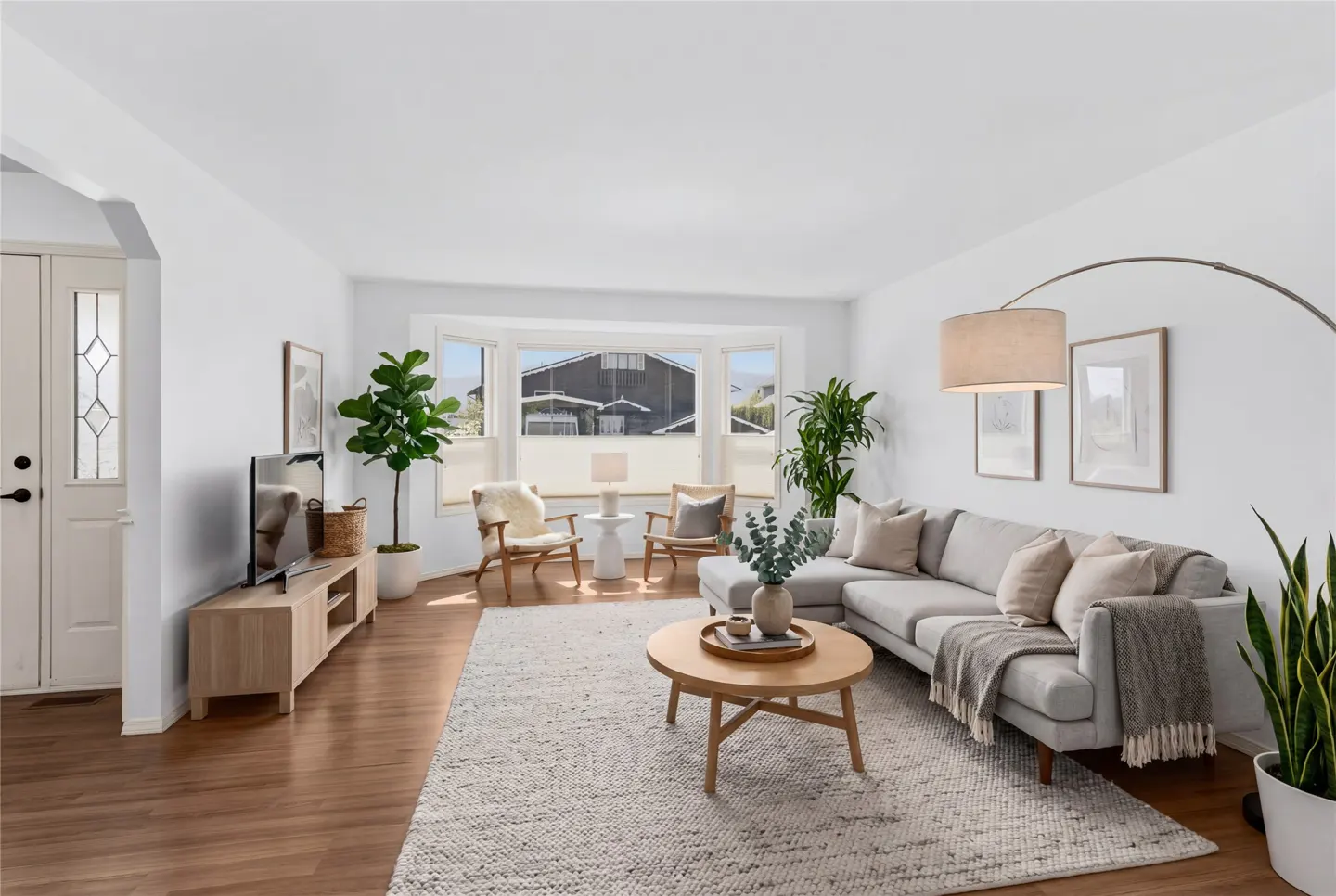 Bright living room with white walls, wood floors, and a gray sectional sofa. A round coffee table sits on a textured rug. Bay window and plants add natural light.