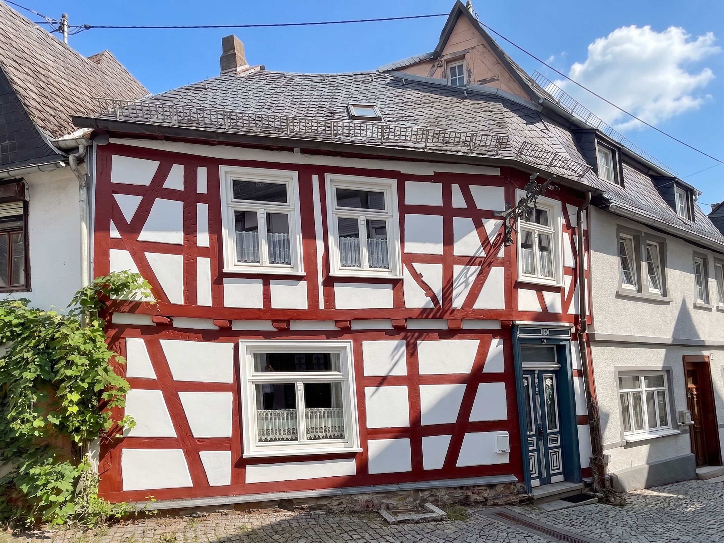 A traditional German half-timbered house with red beams and white walls, featuring a slate roof and a cobblestone street.