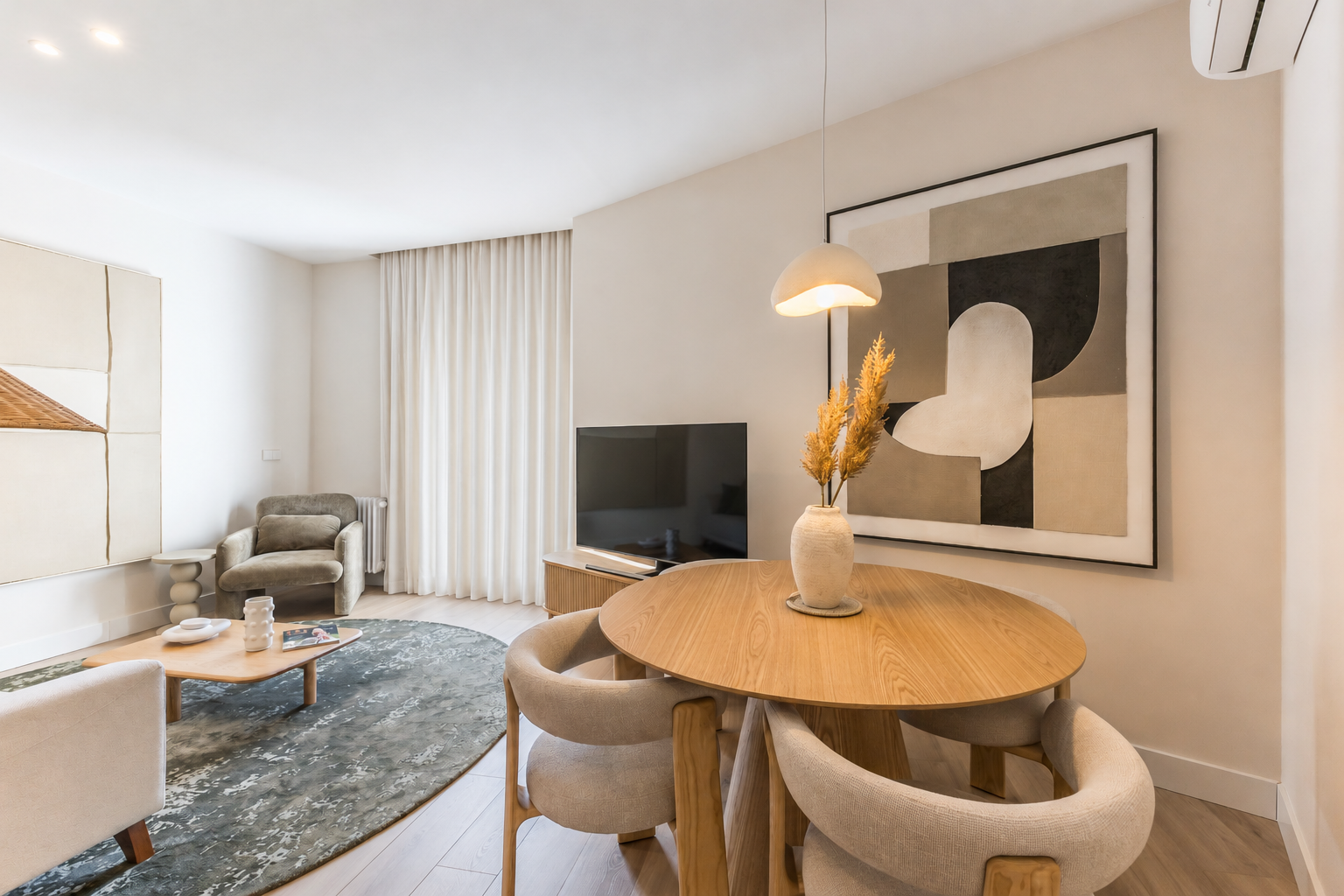 Modern dining area with a round wooden table, beige chairs, and a pendant light, next to a living room with a green rug. [AI-generated text]