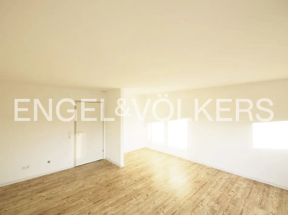 Empty room with light wood-look flooring, white walls, a closed white door, and bright natural light from windows.