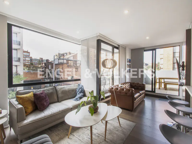 Bright living room with a gray sofa, brown leather couch, and a white coffee table. Large windows offer city views.