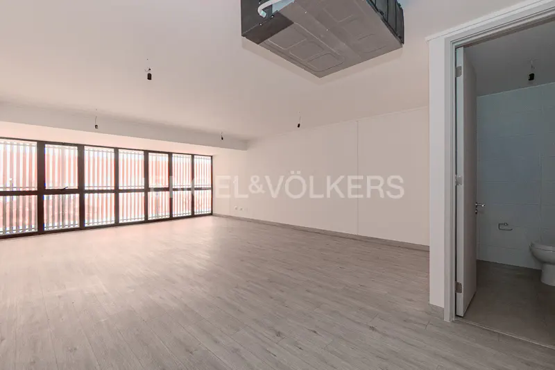 An empty, bright room with light wood floors, a large window with vertical blinds, and an open bathroom door.