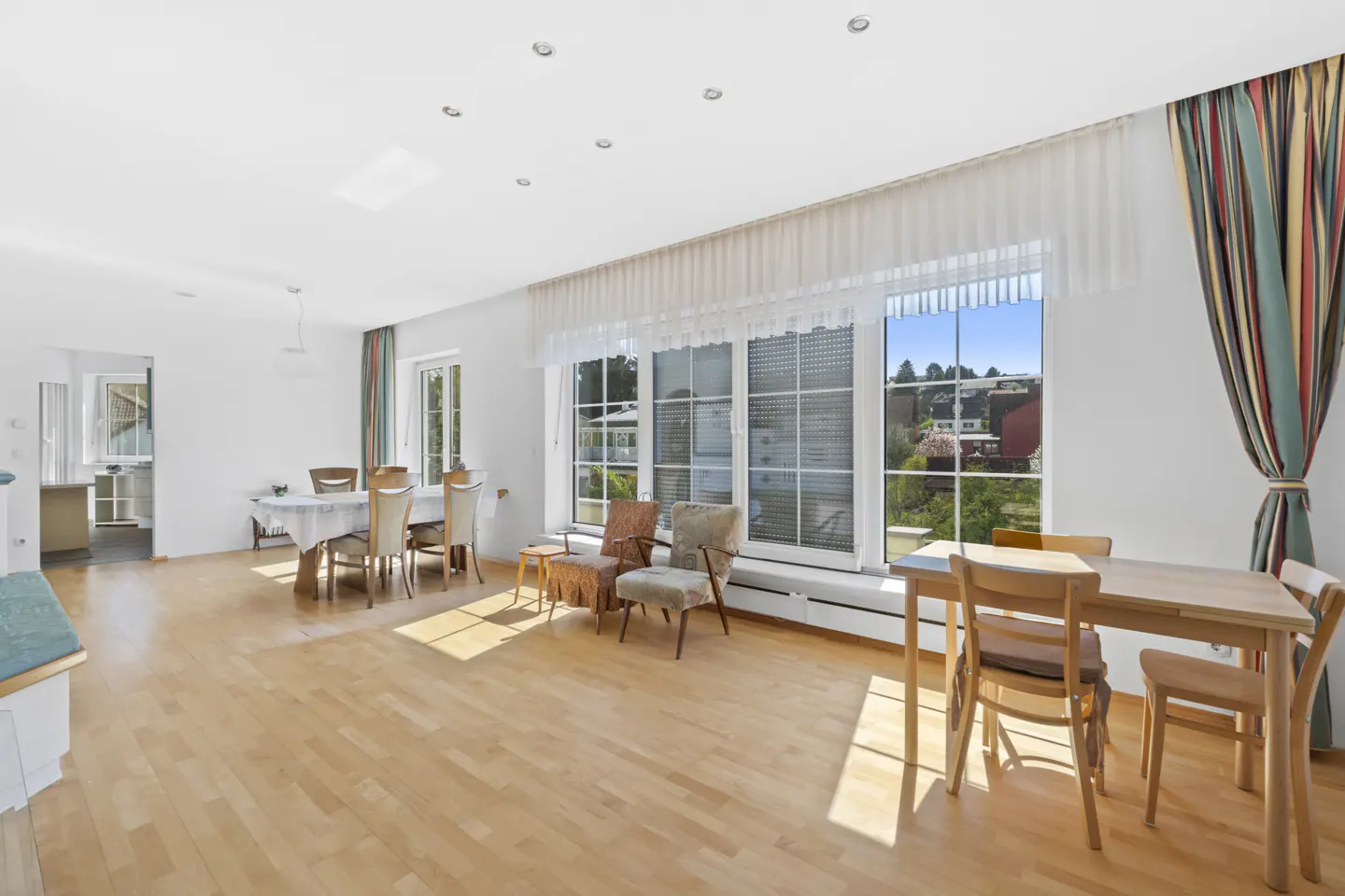 Bright, open living room with wood floors, white walls, and large windows. Dining table, chairs, and striped curtains are visible.