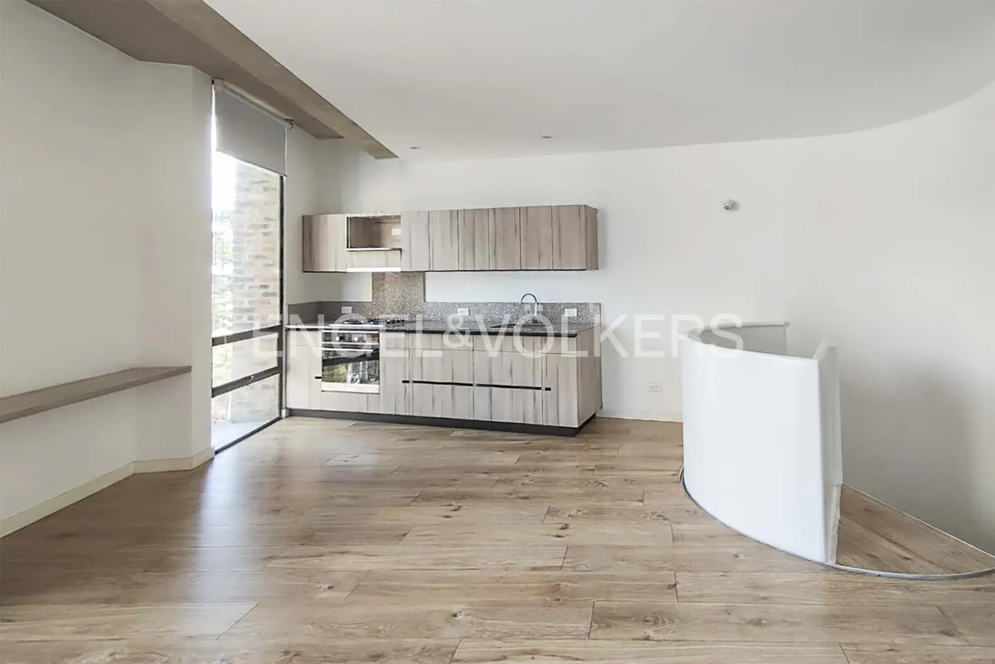 A bright, modern apartment interior with wood floors, a kitchen with light wood cabinets, and a white curved wall.