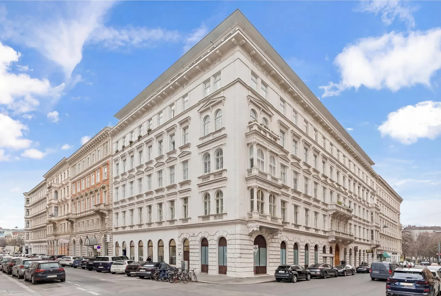Exterior view of a large, white, multi-story building with many windows, parked cars, and a blue sky with clouds.