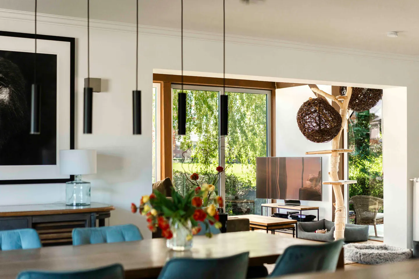 A modern dining room with a wooden table, blue chairs, and black pendant lights. A living room with a TV and cat tree is visible through an opening.