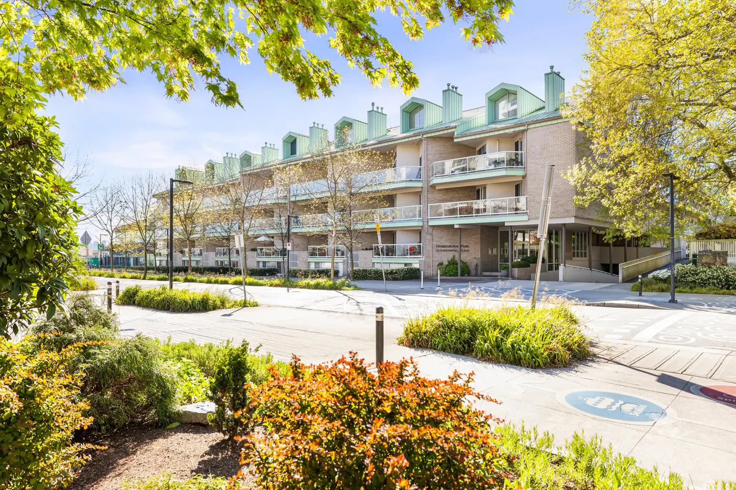 Exterior view of a three-story apartment building with balconies and green roofs, surrounded by trees and landscaping.