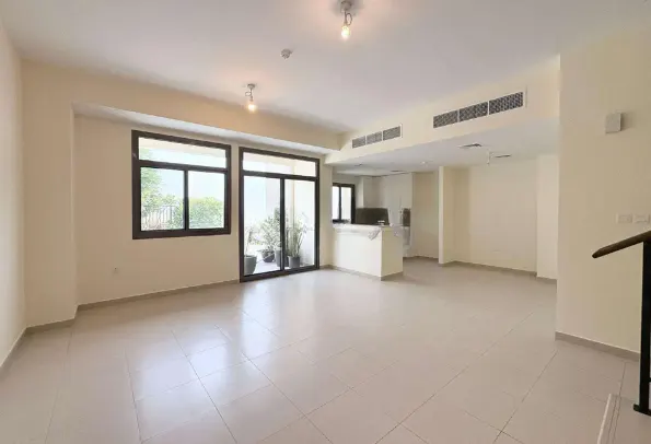 A bright, empty living room with beige walls and tile floors. A sliding glass door leads to a small patio with potted plants.