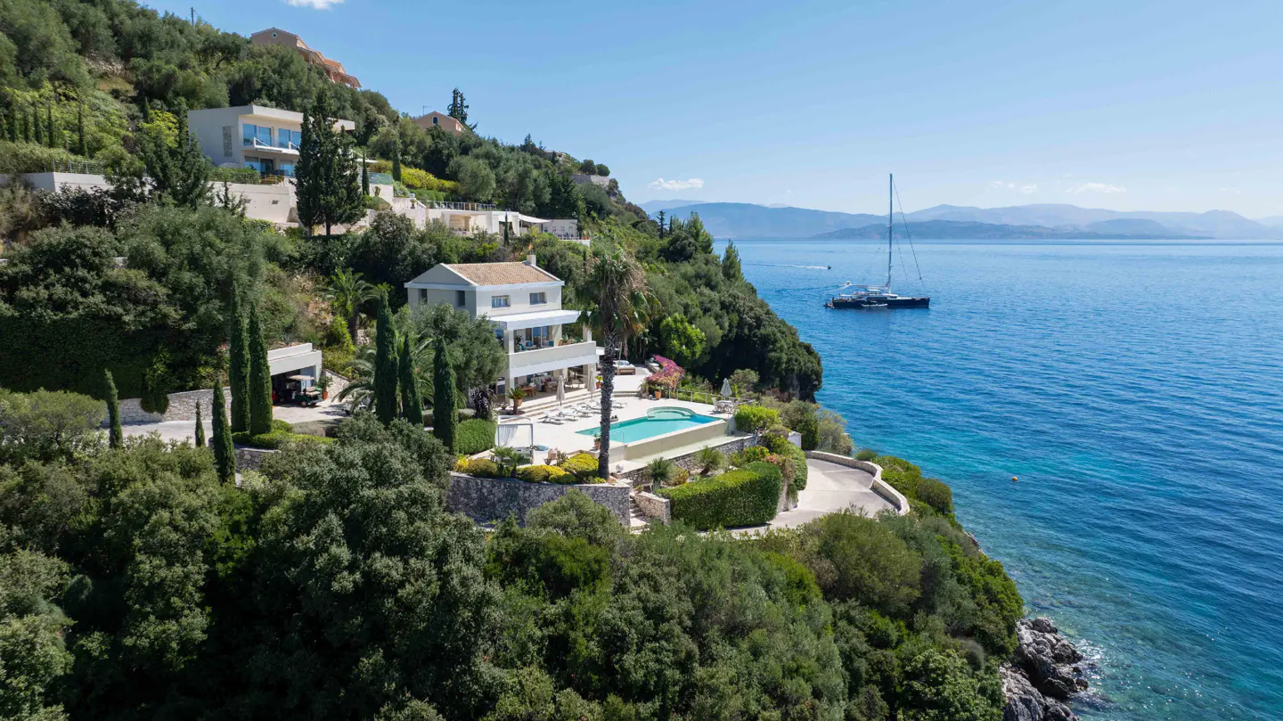 Luxury villa with a pool on a green hillside overlooking the blue ocean with a sailboat.