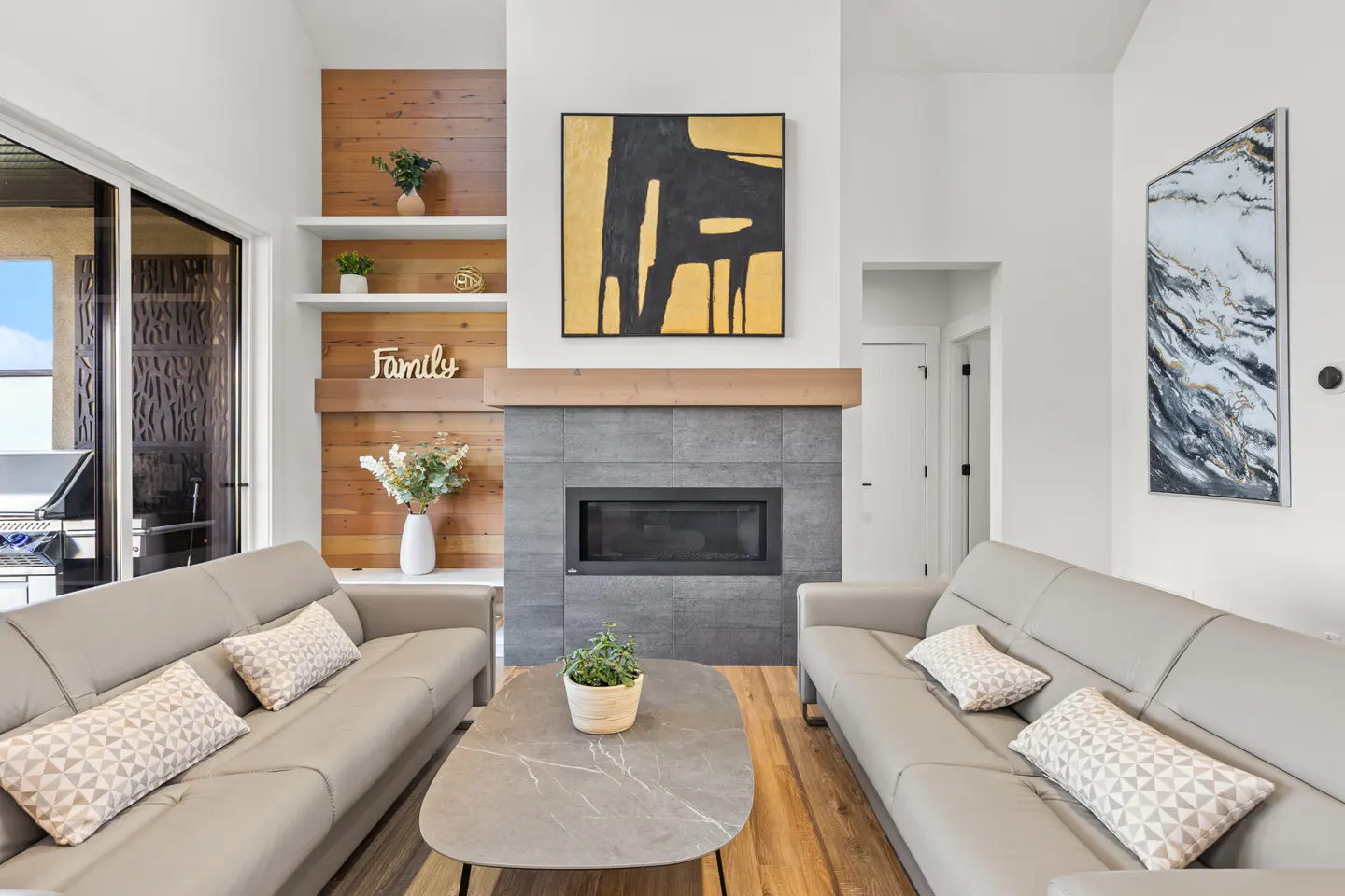 Modern living room with two grey leather sofas, a stone fireplace, wood accent wall, and abstract art.