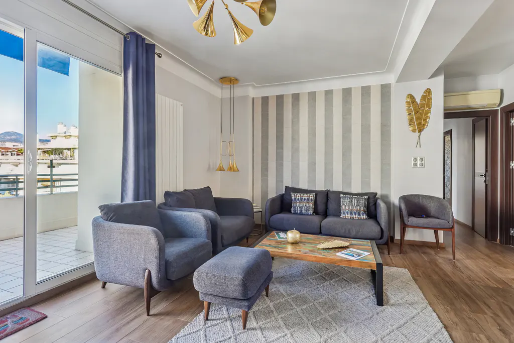 Living room with gray sofa, chairs, and ottoman on a patterned rug. A coffee table sits in the center. Balcony with a view is visible.