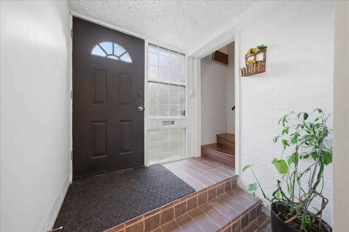 Entryway with a dark brown front door, textured glass sidelight, and brown tiled steps. A potted plant sits near a white brick wall.