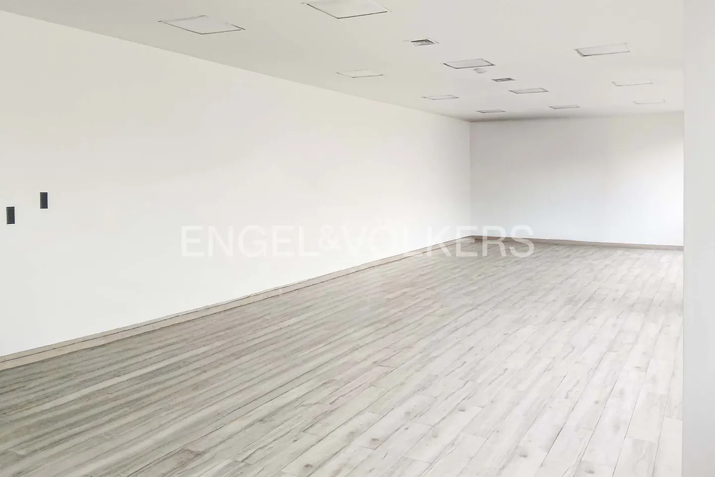Empty room with light wood floors, white walls, and recessed ceiling lights. Engel & Volkers logo on the wall.