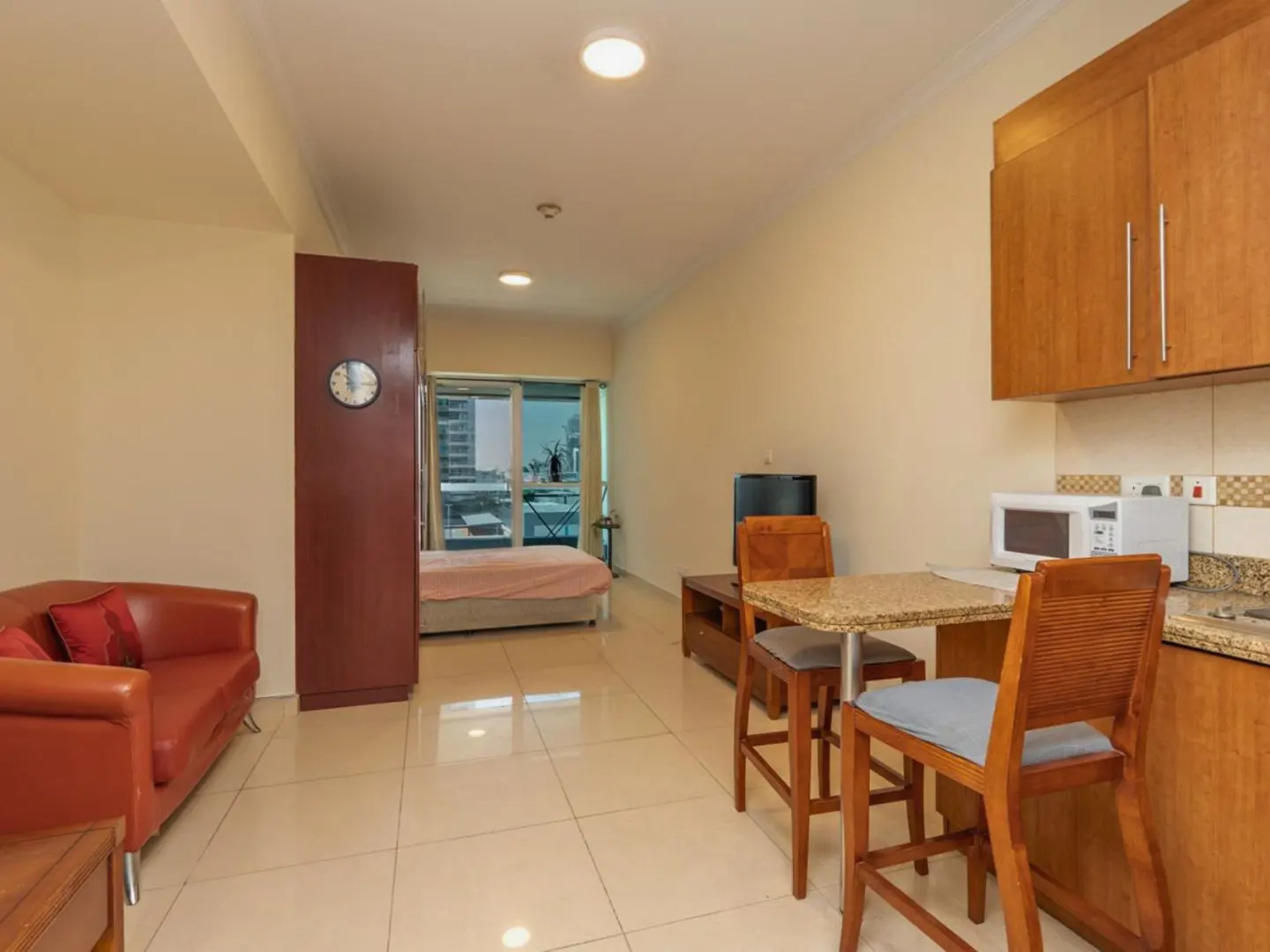 Studio apartment with a red sofa, bed, dining table, and kitchen area. The walls are beige, and the floor is tiled.