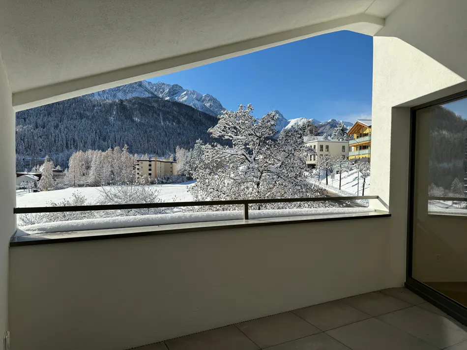 Balcony view of snow-covered mountains, trees, and buildings under a clear blue sky. The balcony has a white wall and a black railing.