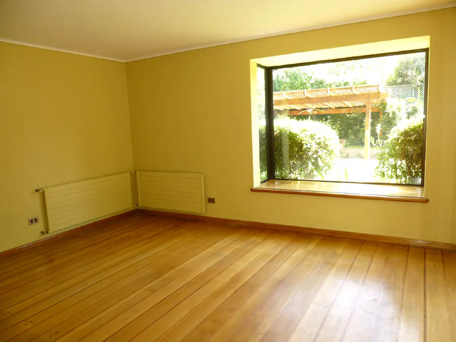 Empty room with light yellow walls, wood floors, and a large window overlooking a garden with a pergola. Radiators are on the wall.