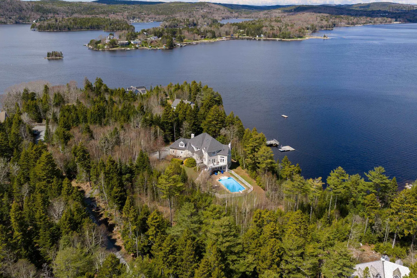 Aerial view of a large, gray house with a pool, surrounded by green trees and a blue lake.