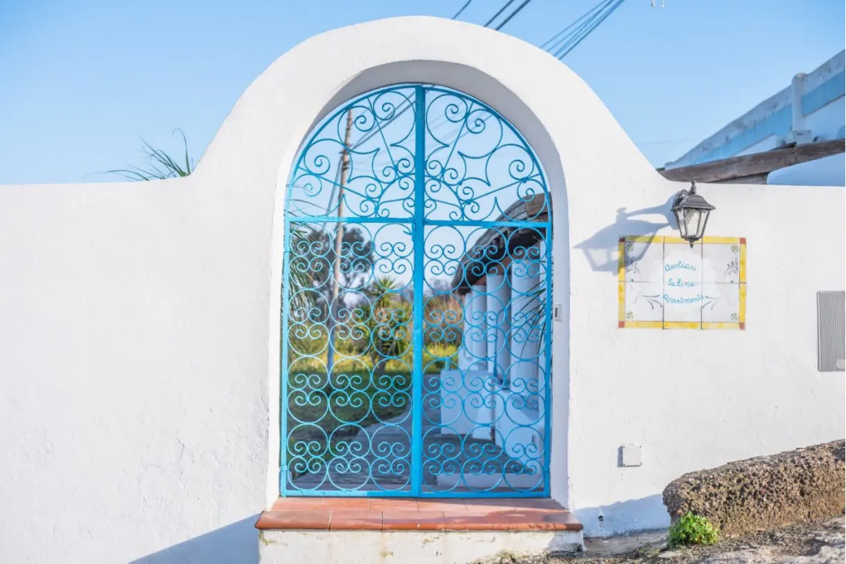 An arched white wall features an ornate, bright blue metal gate leading to a garden, with a decorative tile sign nearby.