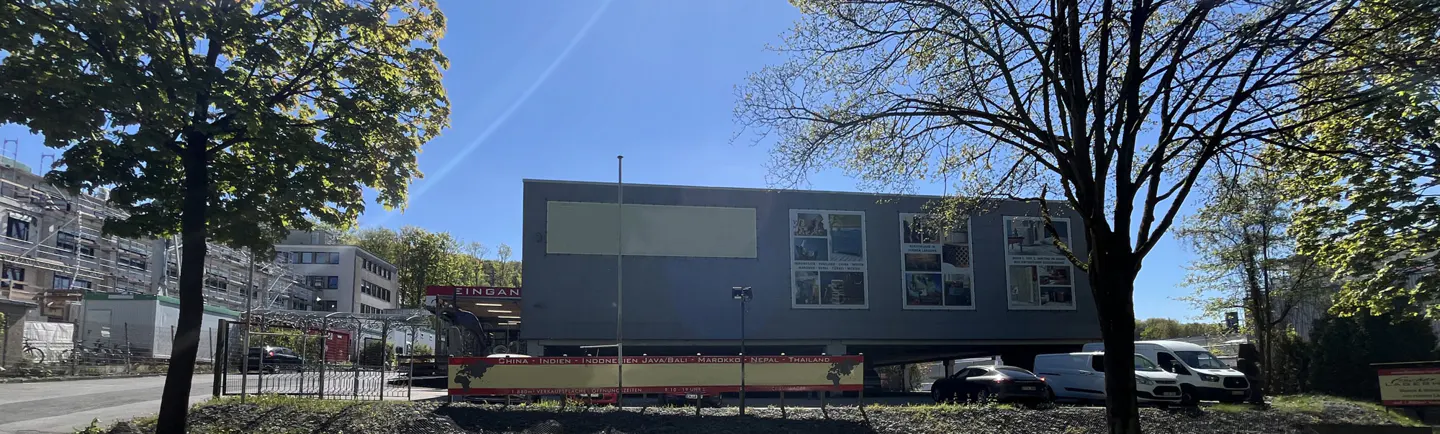 Exterior view of a gray, modern building with "Eingang" sign, surrounded by trees and parked vans under a sunny, blue sky.