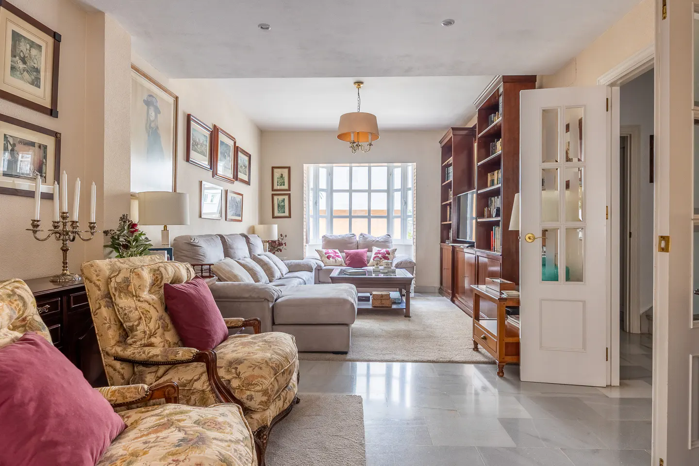 Bright living room with floral armchairs, a gray sectional sofa, and a dark wood bookcase. Artwork adorns the walls.