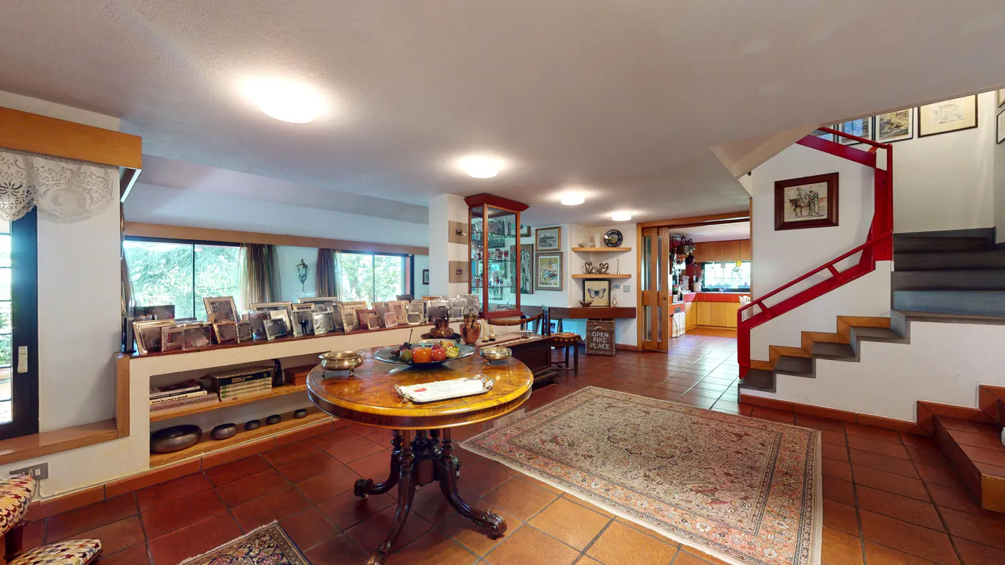 A bright foyer with terracotta tile floors, a round wooden table, and a staircase with red railings. Shelves display photos and books.