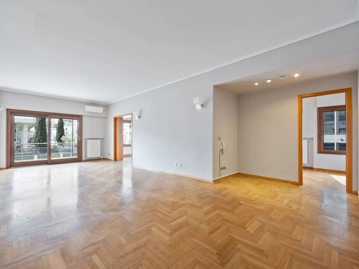 Bright, empty room with herringbone wood floors, gray walls, and white ceiling. Sliding glass doors lead to a balcony.