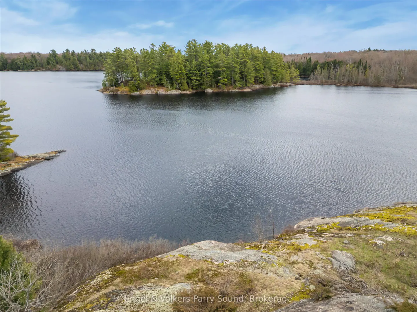 Scenic view of a calm lake with a tree-covered island, seen from a rocky, mossy overlook under a blue sky.
