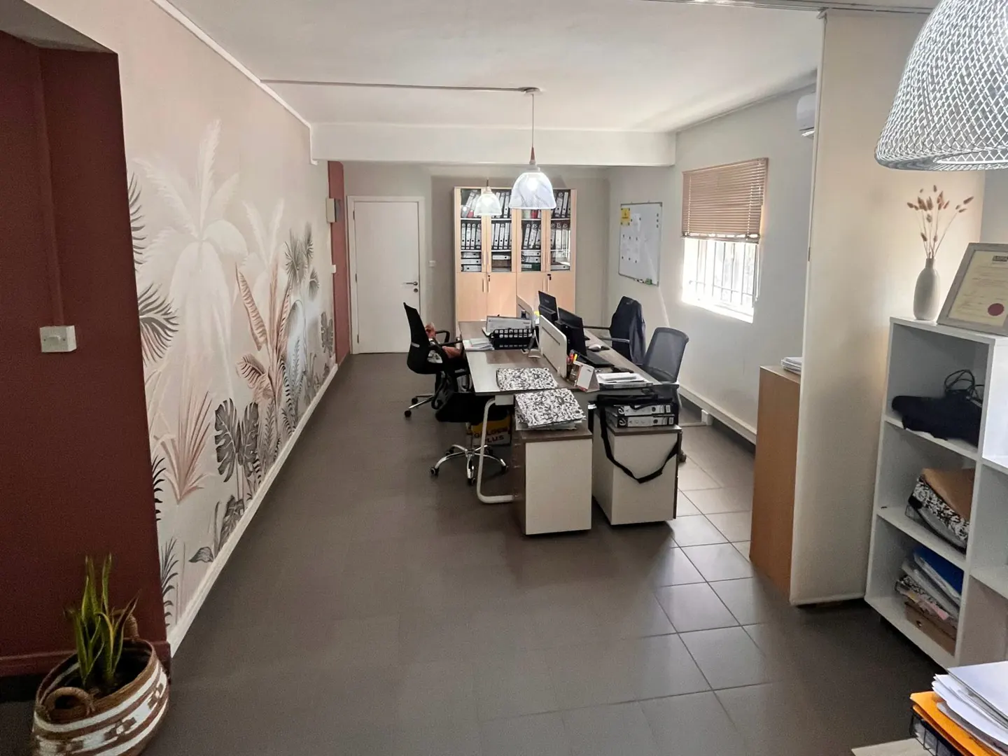 Modern office with a tropical-themed mural, long desk with chairs, storage cabinets, and a white shelving unit.