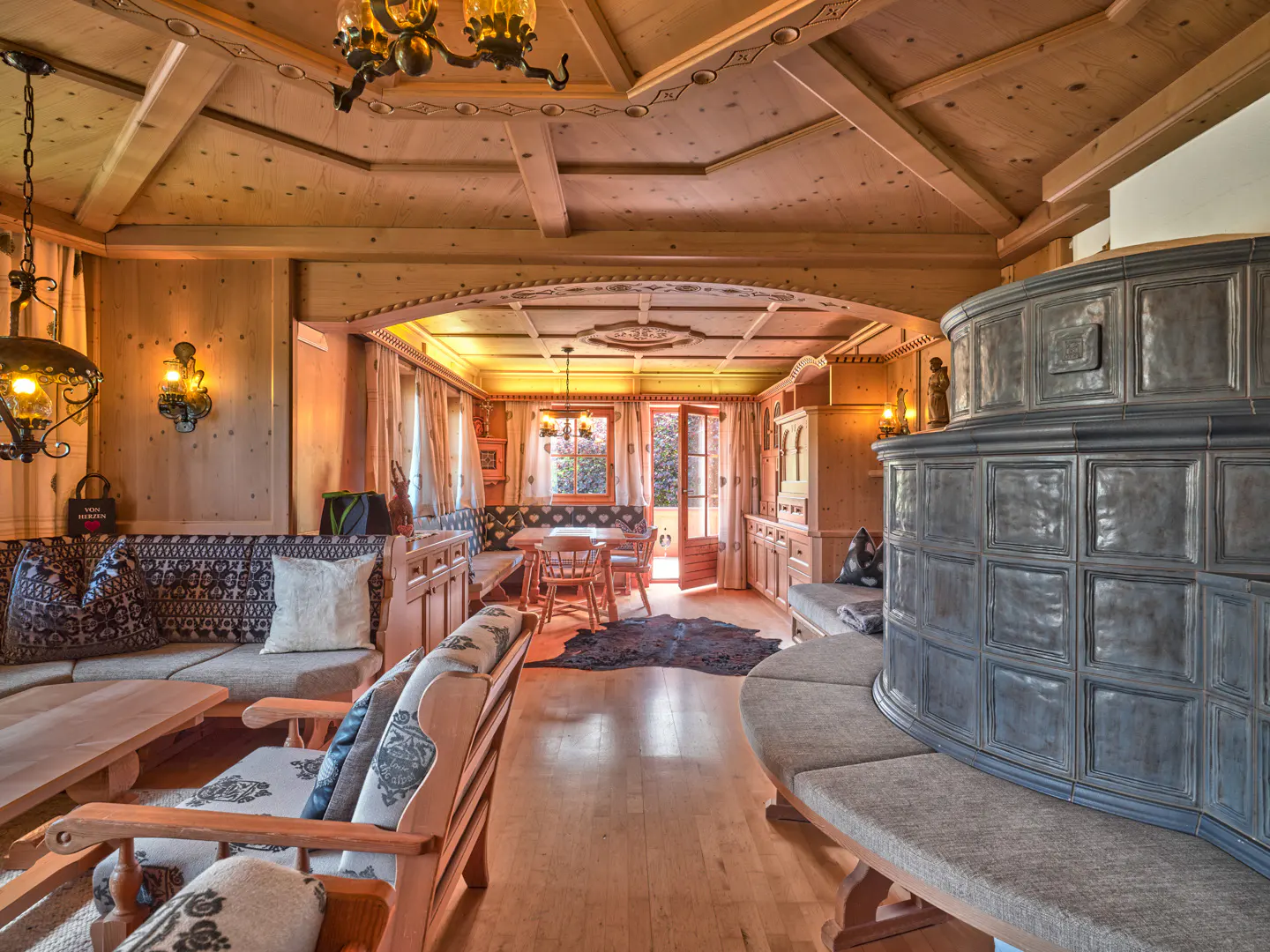Rustic living room with wood-paneled walls, a large grey tiled stove, patterned seating, and a dining area in the back.
