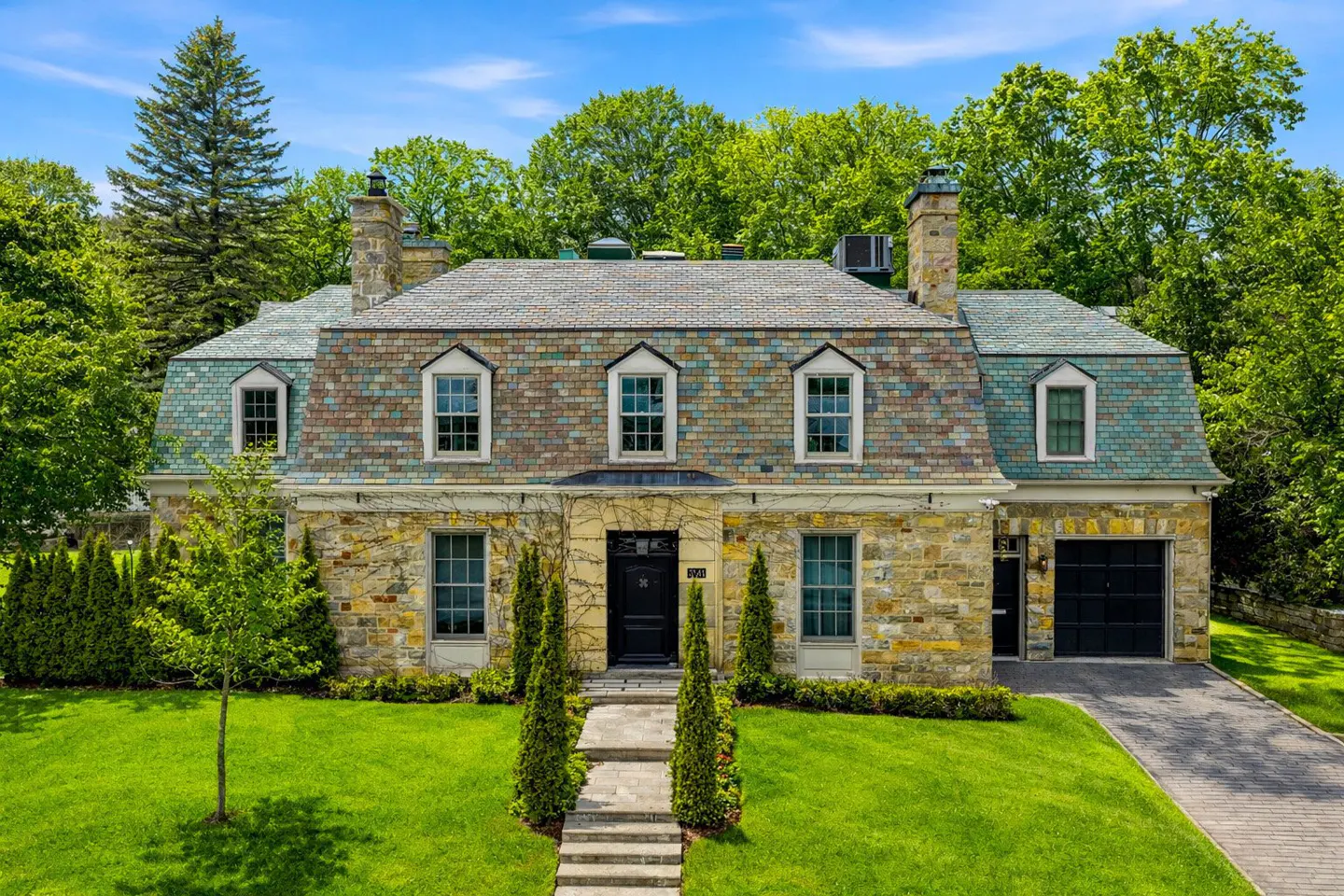 Stone house with a gray and teal roof, black door and garage, green lawn, and trees under a blue sky.