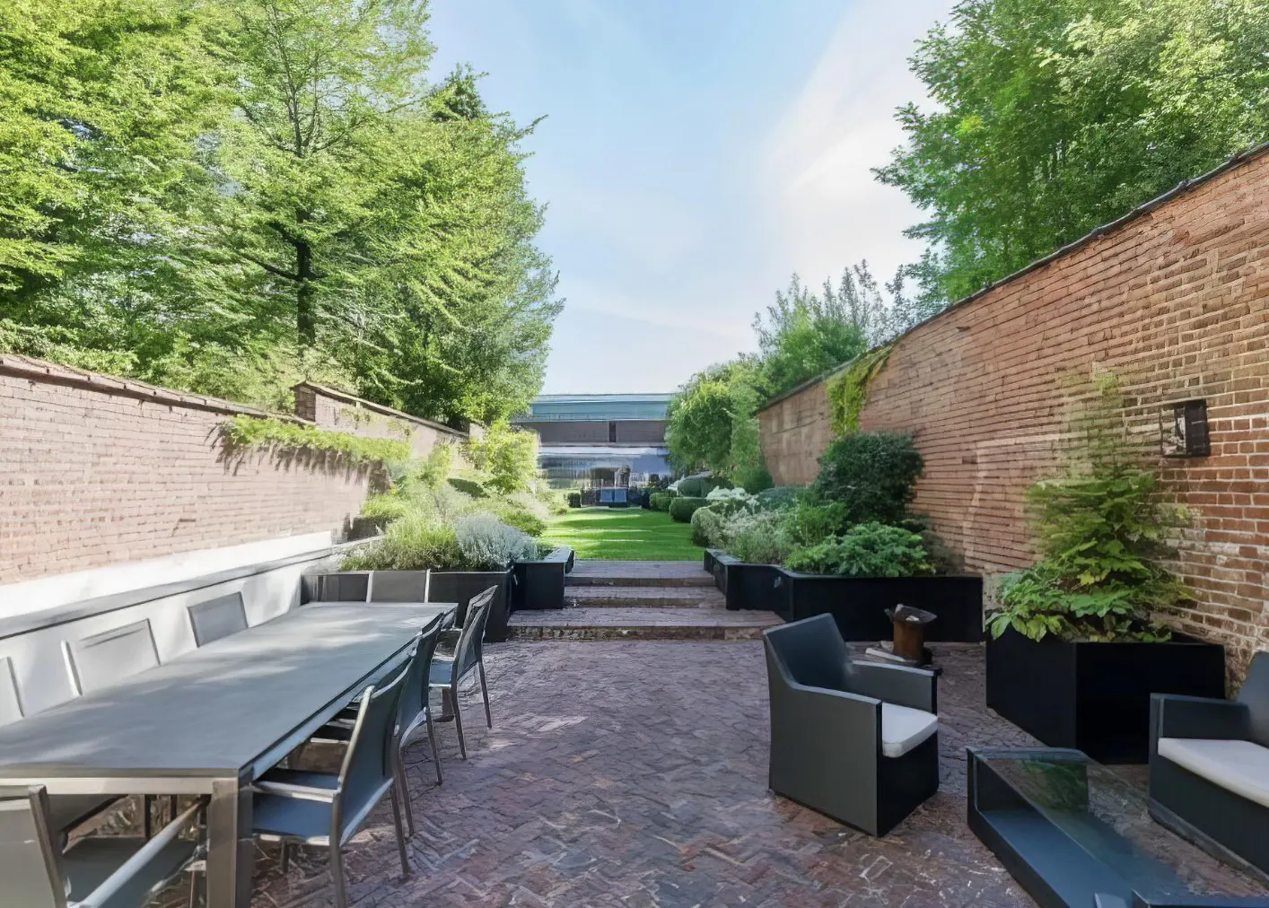 Outdoor patio with brick flooring, brick walls, green trees, and outdoor furniture including a long table and chairs.