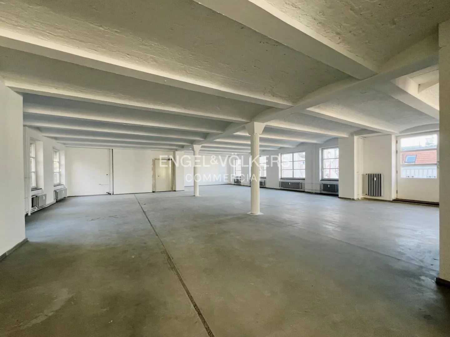 An empty commercial space with white walls, concrete floors, and large windows. White support columns and exposed ceiling beams are visible.