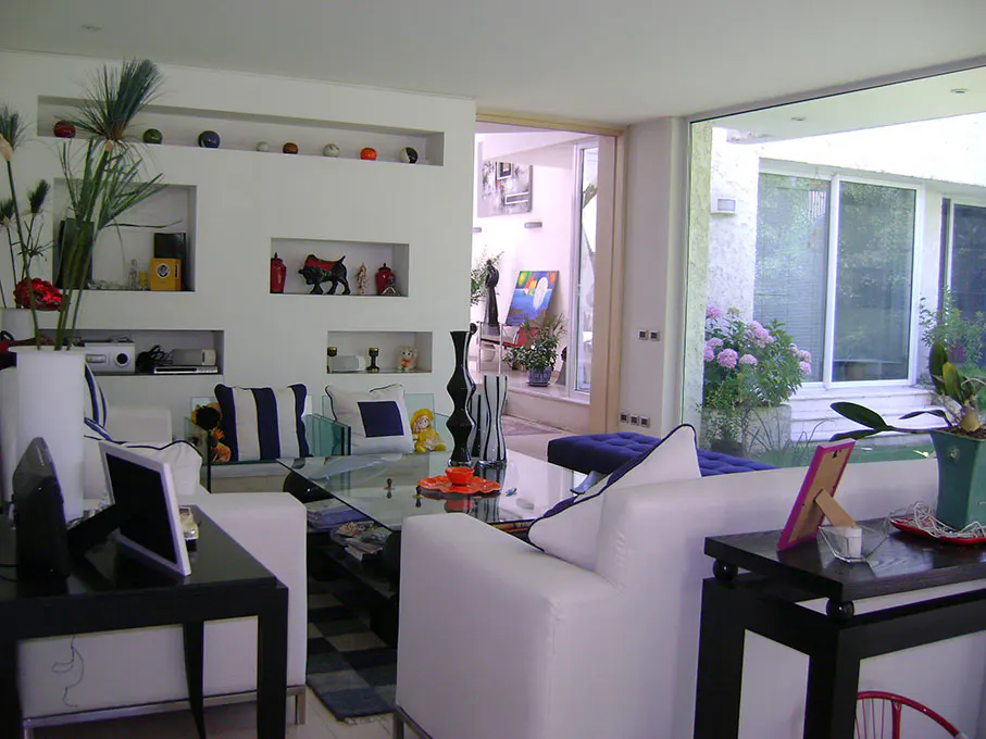 Bright living room with white sofas, a glass table, and black side tables. Built-in shelves display decorative items. Large windows offer a view of the garden.