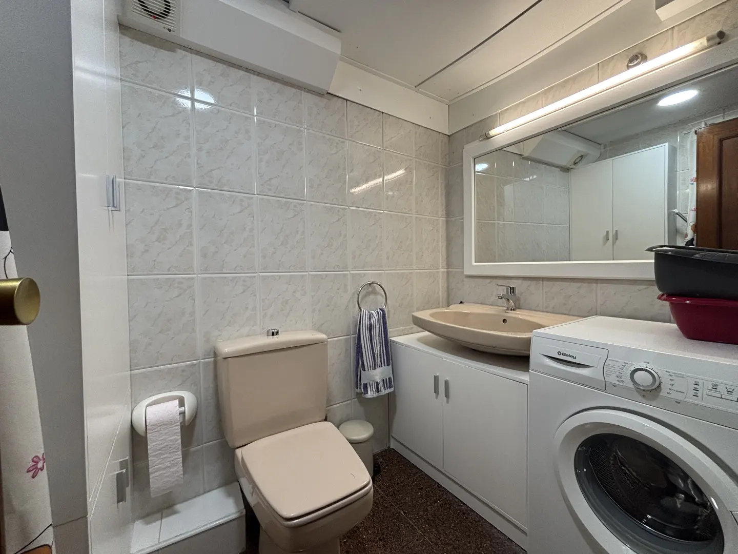A small bathroom with a beige toilet, sink, and white washing machine. The walls are tiled in a light pattern. A mirror hangs above the sink.