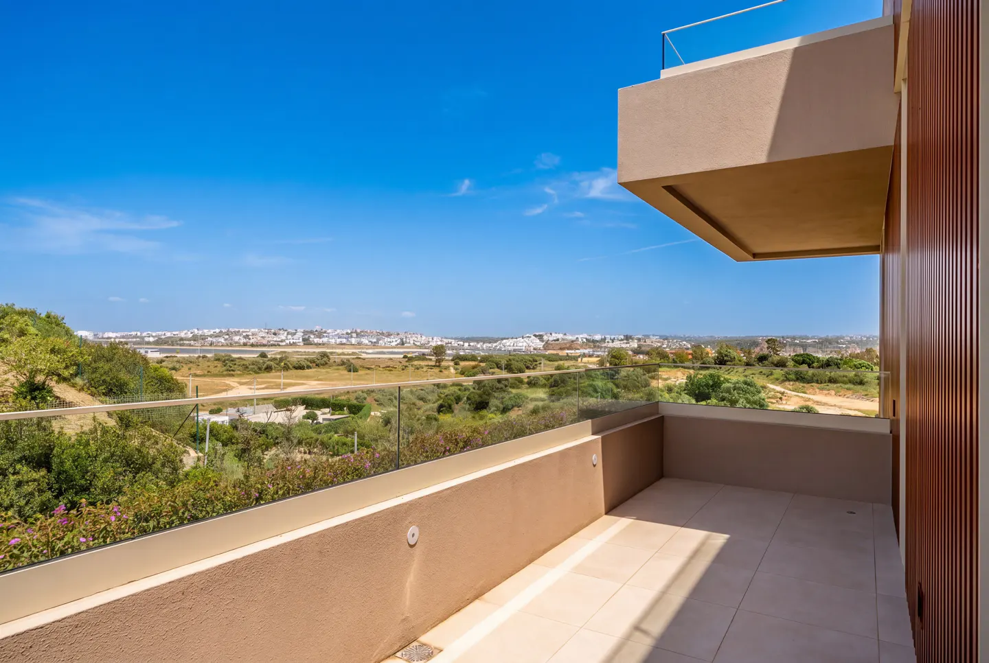 Balcony view with glass railing overlooking green landscape and distant city under a clear blue sky. Beige walls and tile flooring.