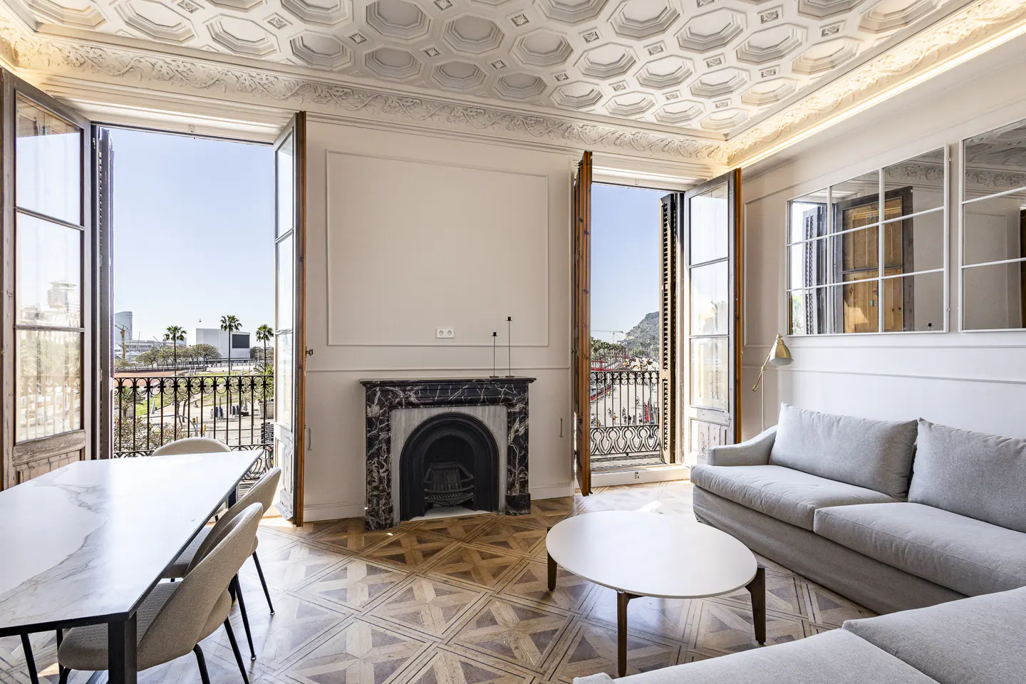 Bright living room with open French doors to balconies. Marble fireplace, patterned wood floor, and gray sofa.