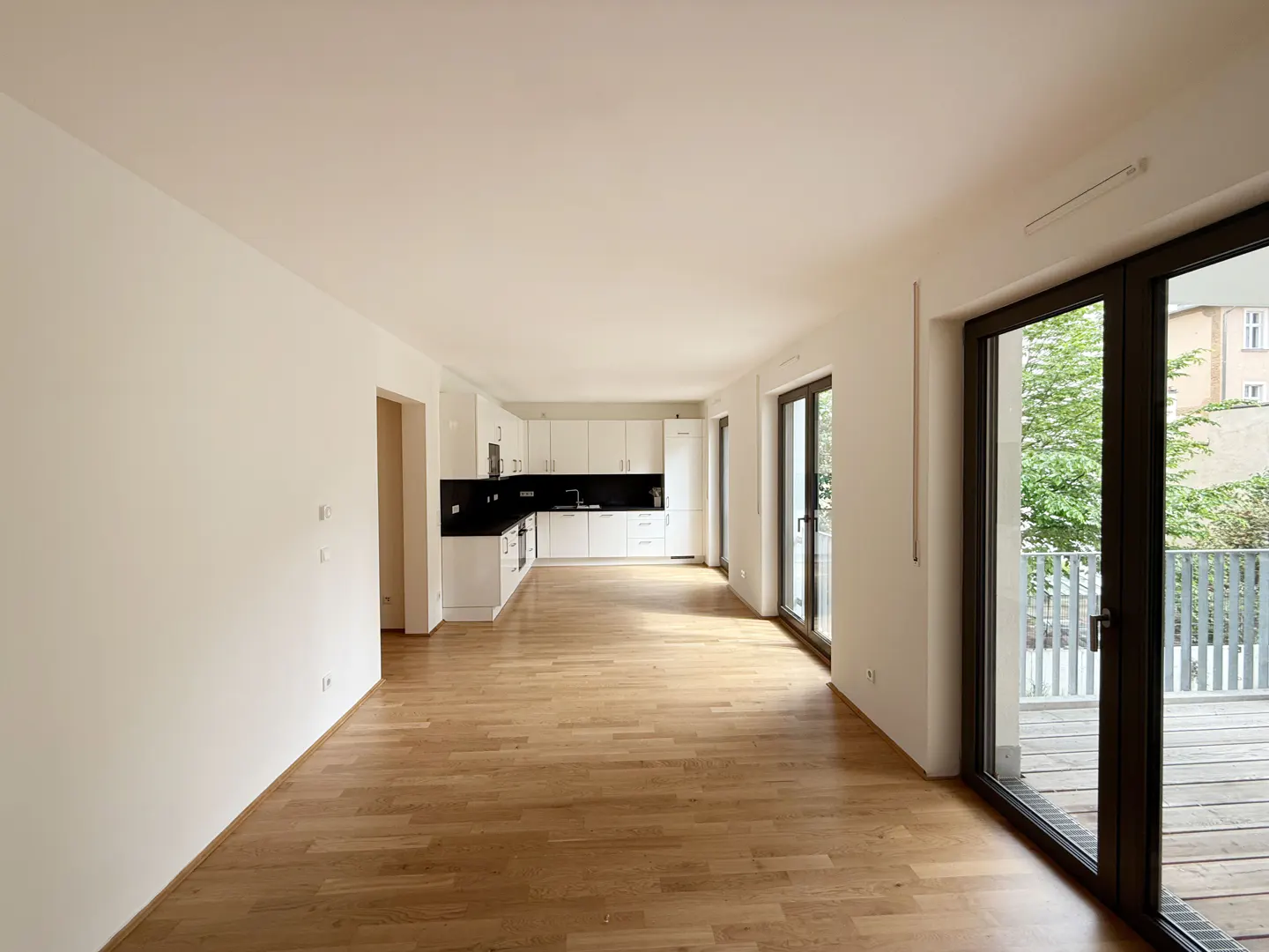 Bright, empty apartment with hardwood floors, white walls, and a modern white kitchen with black countertops. Balcony access through sliding glass doors.