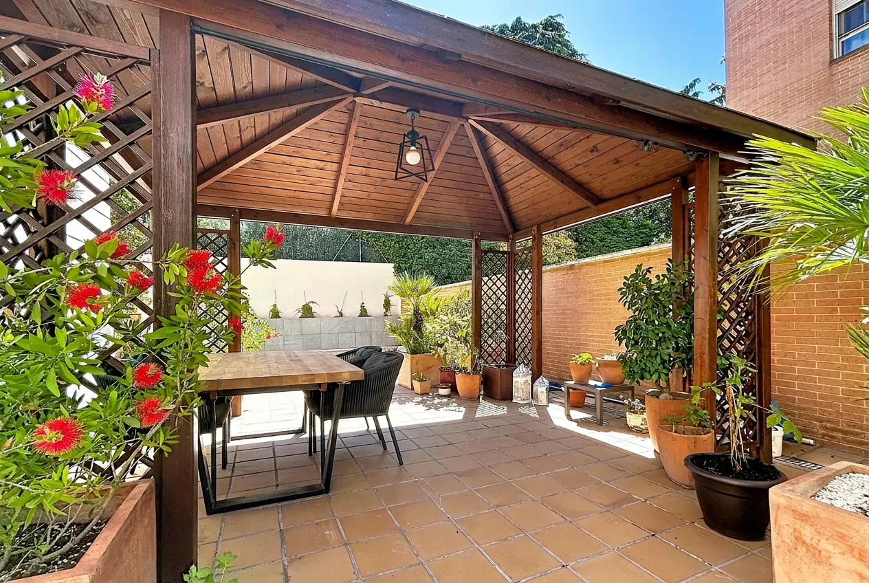 A backyard patio features a wooden gazebo with a table and chairs, surrounded by potted plants and flowers.