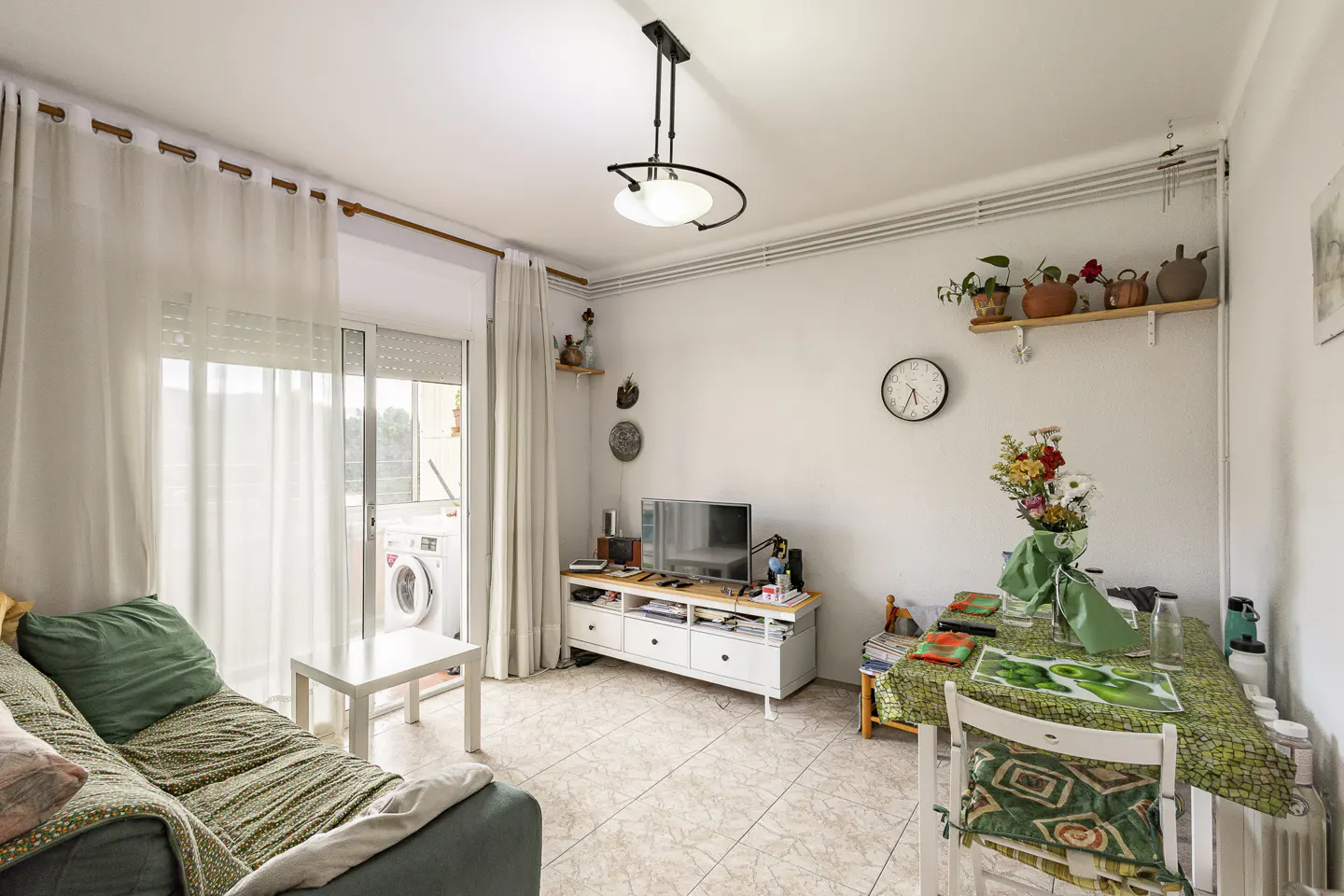 Bright living room with a green sofa, white curtains, and a table with flowers. A TV and shelves decorate the white walls.