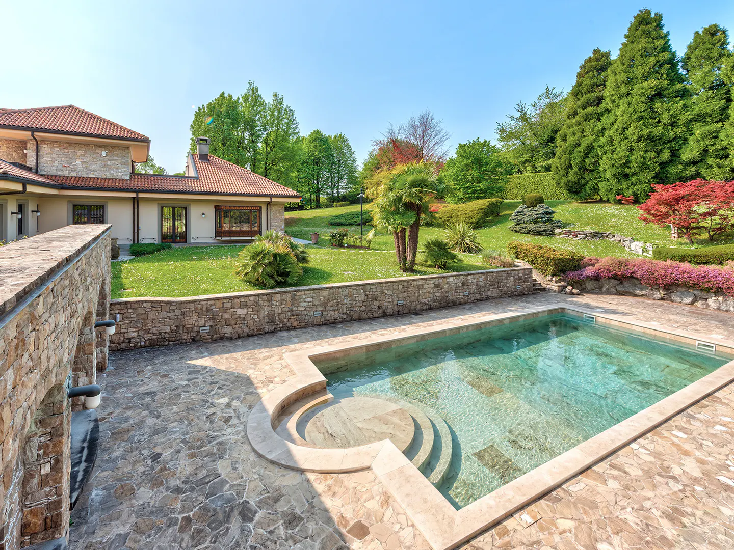 Outdoor pool with stone patio and steps, surrounded by green lawn, trees, and a stone house under a blue sky.