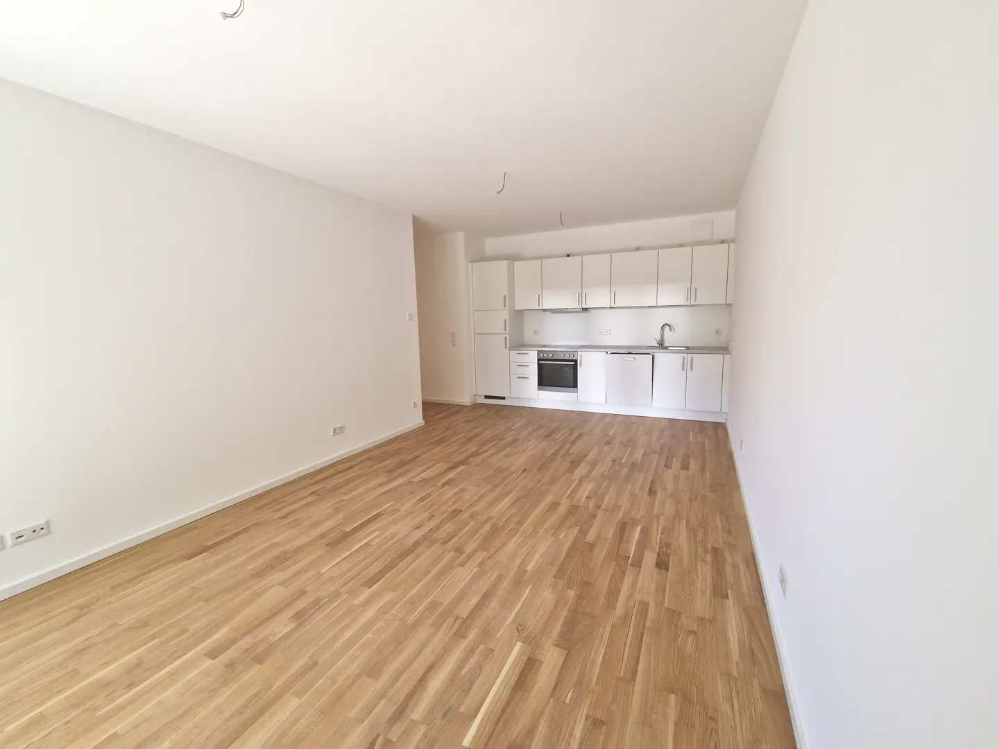 Bright, empty apartment with light wood floors and white walls. A white kitchen is in the background.