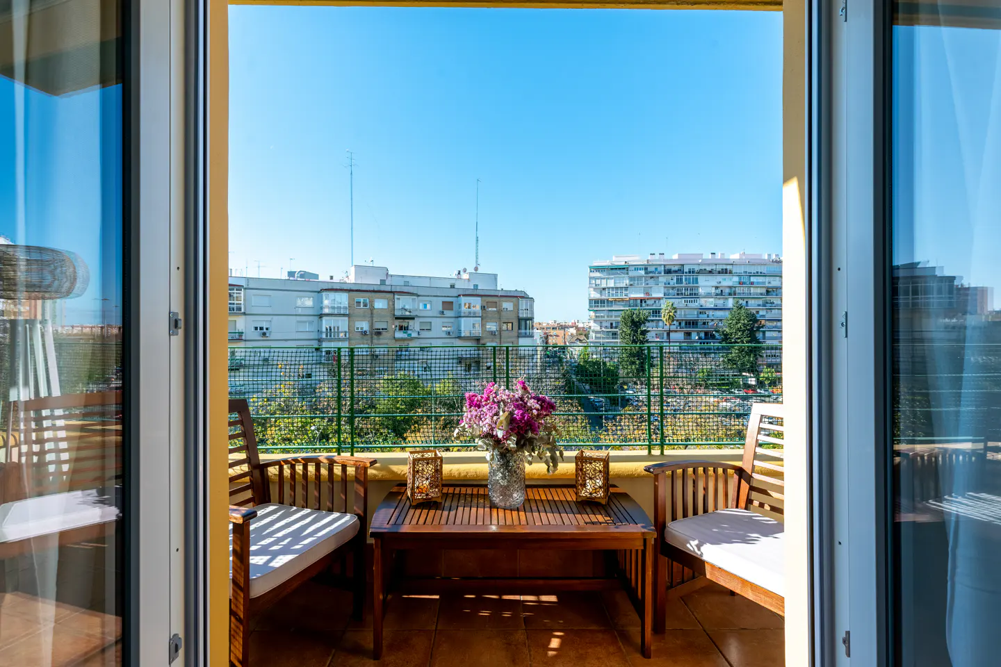 Balcony view with wooden chairs, table, and flowers. Buildings and blue sky in the background.