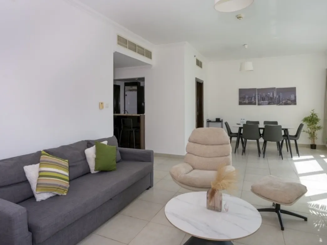 Bright living room with gray sofa, beige chair and ottoman, and a white marble table. A dining table with six chairs is in the background.