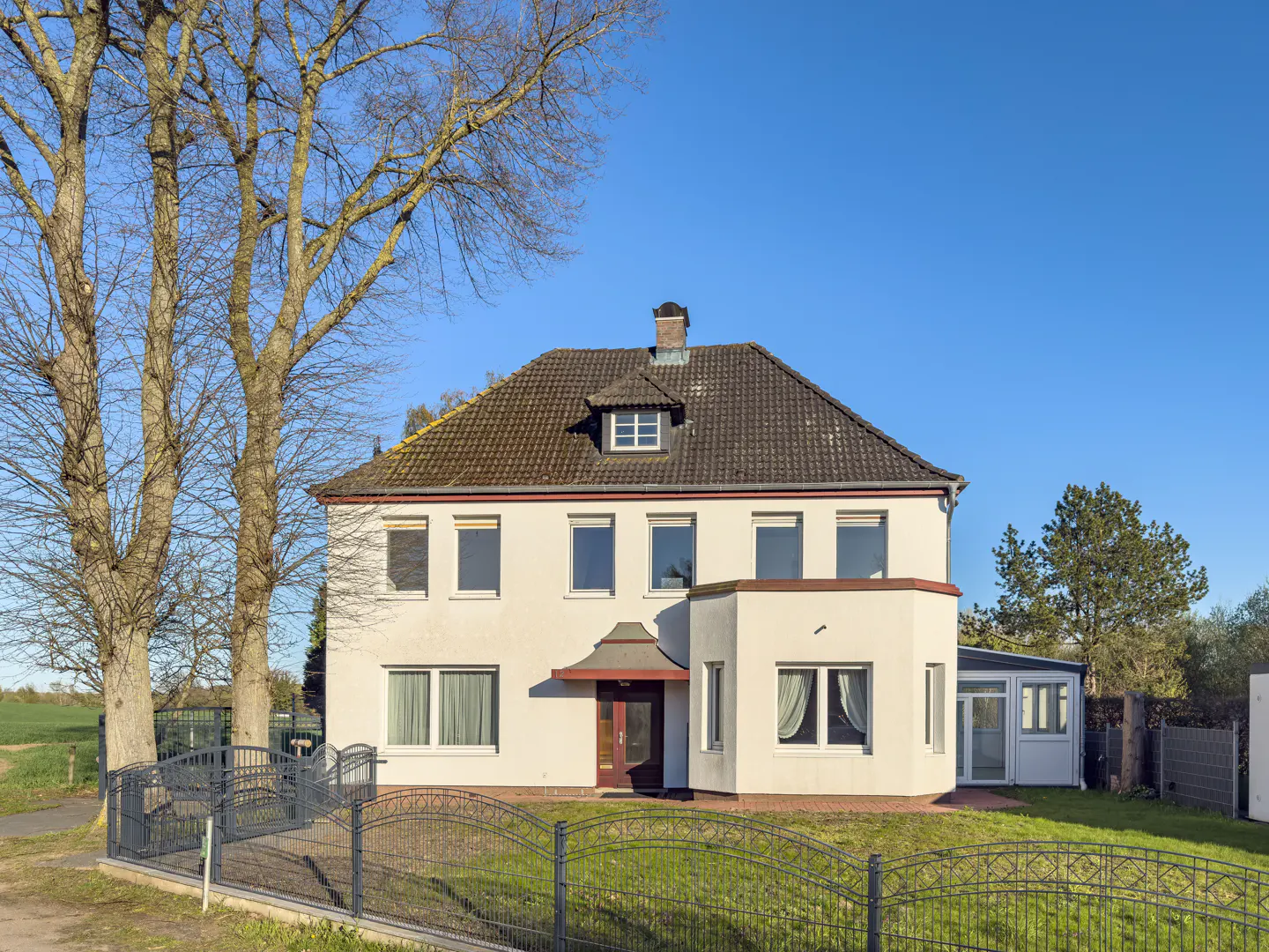 Two-story white house with a brown roof, chimney, and dormer window, surrounded by a gray fence, grass, and trees under a blue sky.