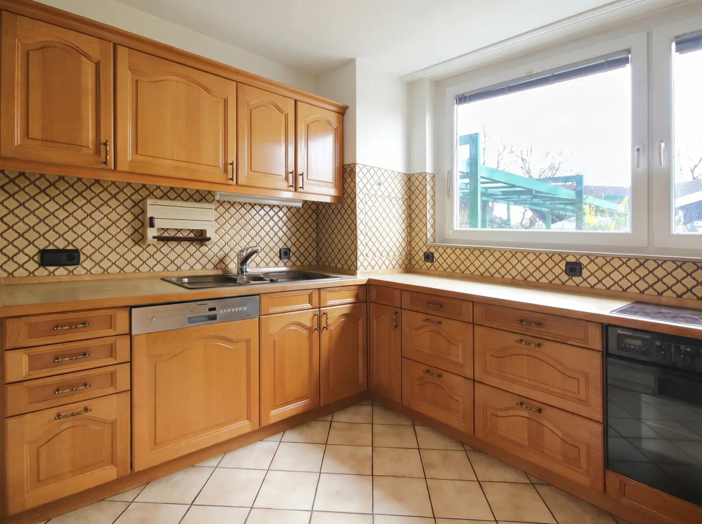 A kitchen with light brown cabinets, beige countertops, and diamond-patterned backsplash tiles. A window overlooks a green structure outside.