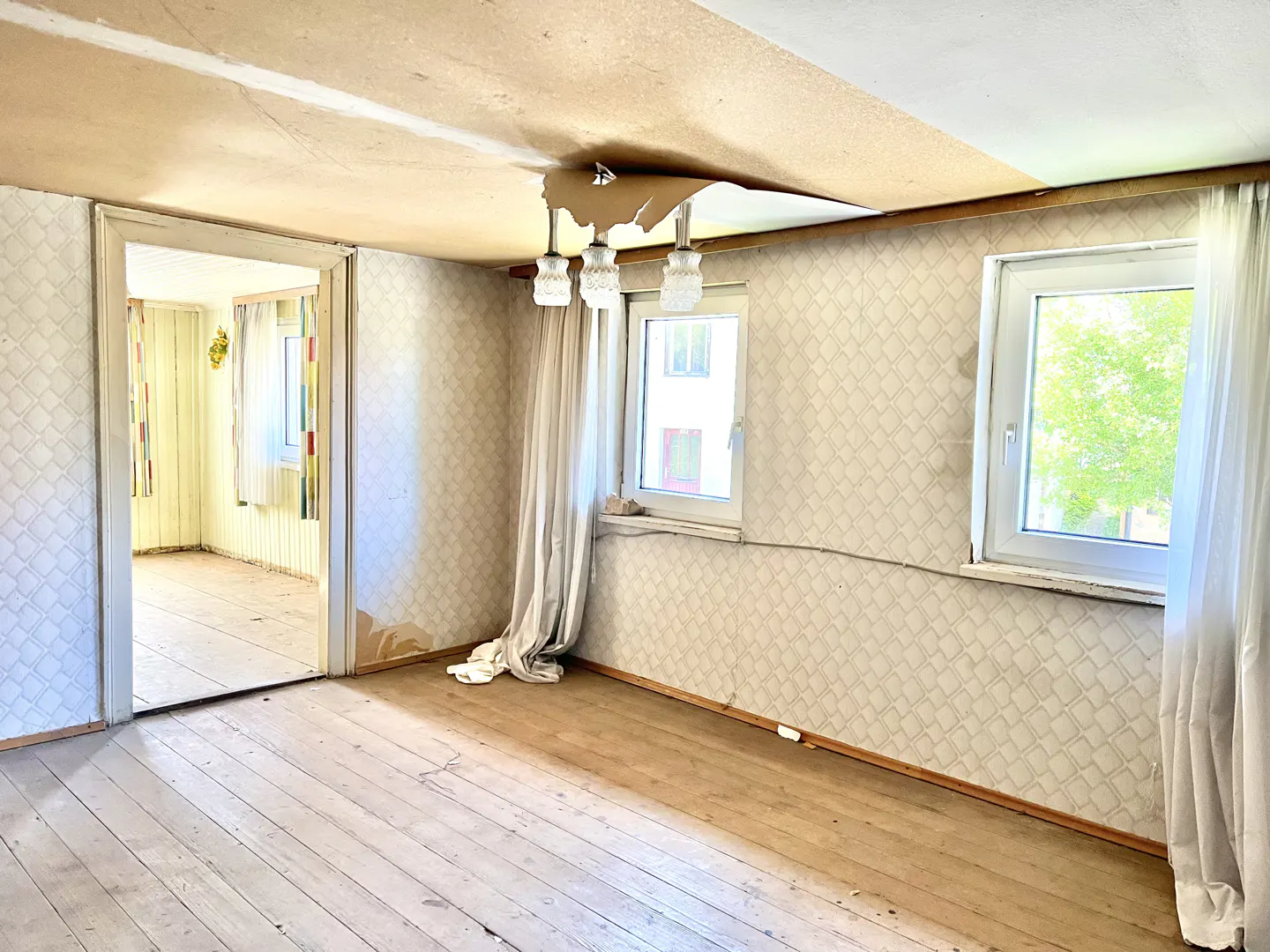 A room with peeling ceiling paint, patterned wallpaper, and two windows with white curtains. A doorway leads to another room with yellow walls.