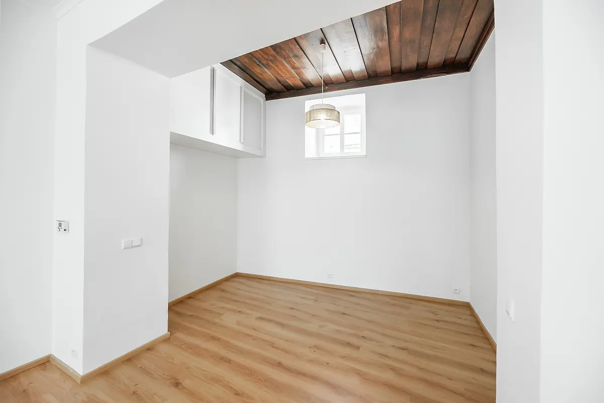Bright, empty room with wood floors, white walls, and a dark wood ceiling. A window and light fixture are visible.