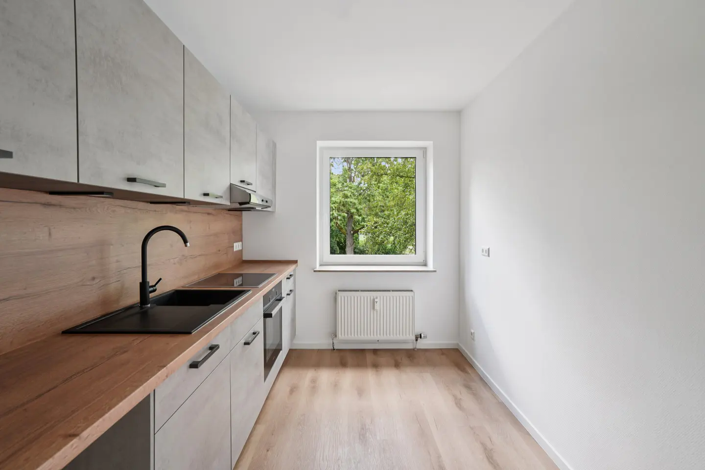Modern galley kitchen with light grey cabinets, wood countertops, black sink, and a window overlooking green trees.