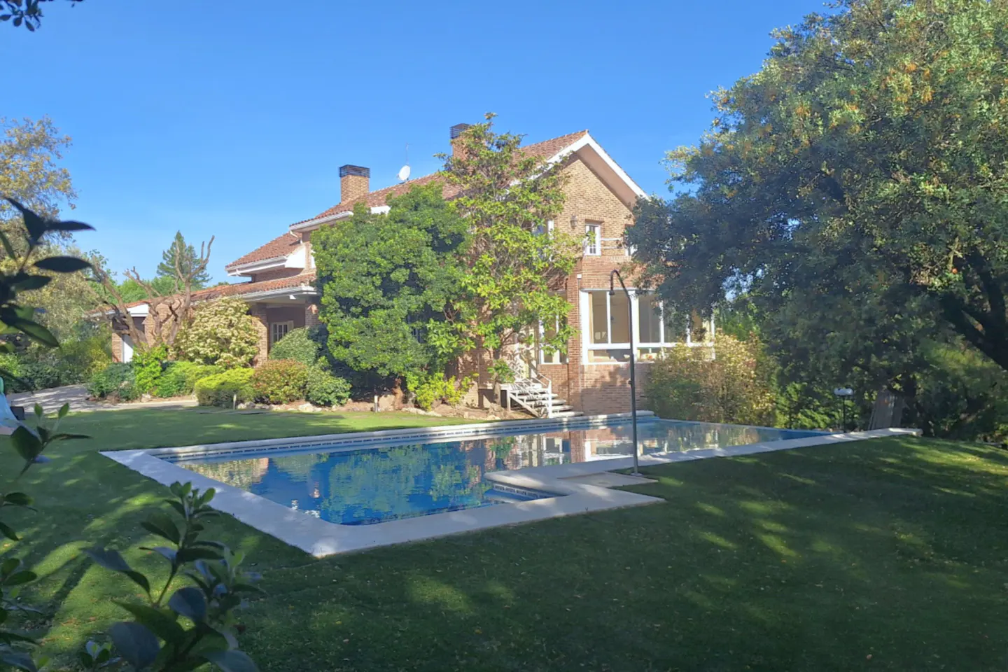 Brick house with a red tile roof, surrounded by green trees and grass, with a blue swimming pool in the foreground.
